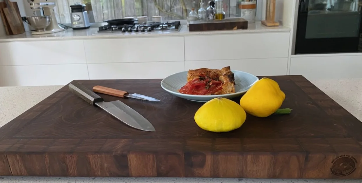 Handcrafted walnut cutting board in a working kitchen with knives, squash, and a prepared dish